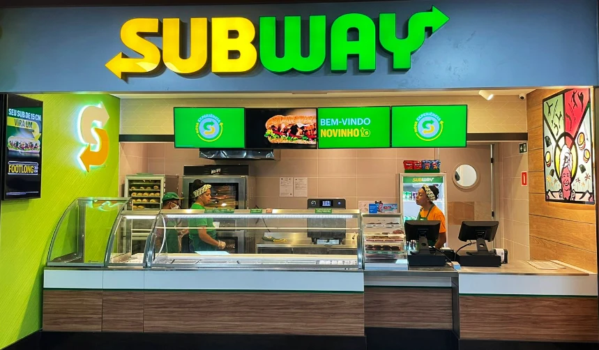 Balcão de atendimento de uma loja Subway, com vitrine de ingredientes, dois funcionários e letreiro SUBWAY acima.