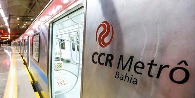 Trem parado em uma estação, com a porta aberta e o nome “CCR Metrô Bahia” visível na lateral.